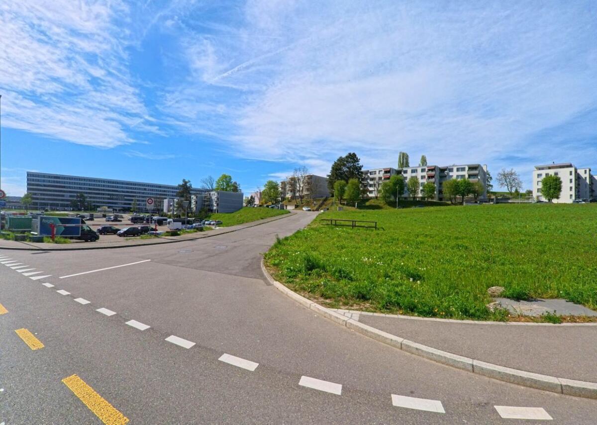 Am Stadtrand von Opfikon (8152, Kanton Zürich) grenzen moderne Wohnanlagen an eine grosse Gruenflaeche neben einer Strasse mit Parkplatz.