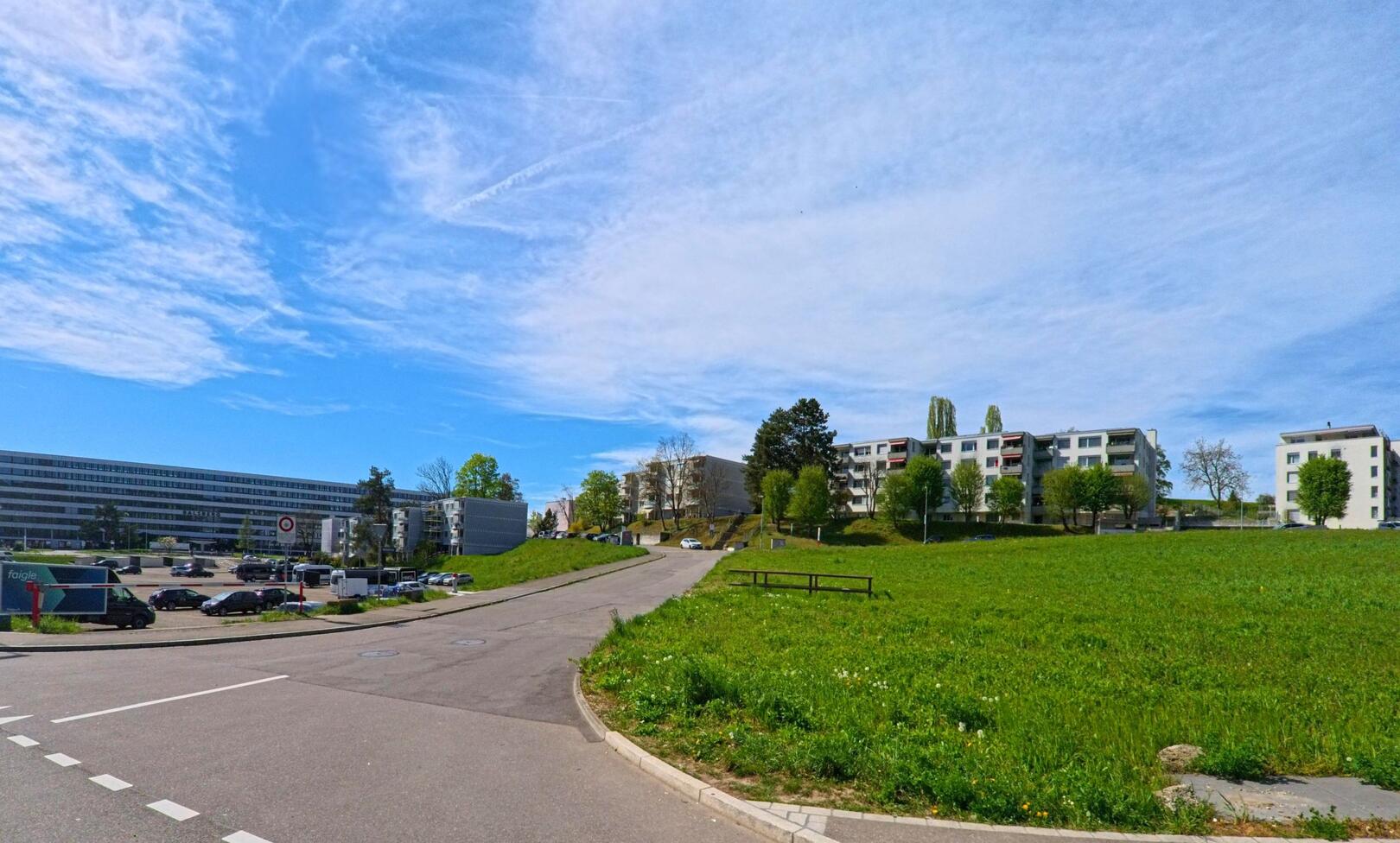 In Opfikon (8302, Kanton Zürich) steigt eine Wohnstrasse zwischen modernen Mehrfamilienhäusern sanft den Hügel hinauf, flankiert von einer grossen Grünfläche und einem Parkplatz.