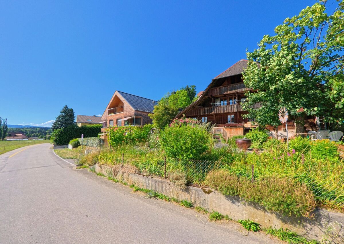 In Oppligen (3629, Kanton Bern) stehen ein traditionelles Bauernhaus mit Holzfassade und ein modernes Gebäude mit Solarpanels nebeneinander an einer ruhigen Landstrasse.