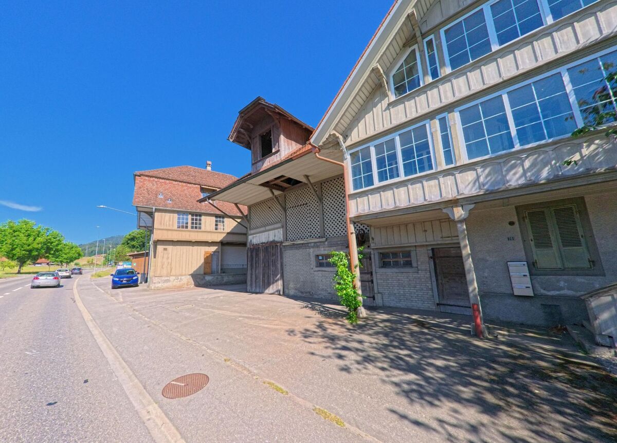 In Oppligen (3629, Kanton Bern) stehen mehrere traditionelle Gebäude mit markanten Dachformen und grossen Fensterfronten direkt an einer vielbefahrenen Hauptstrasse.