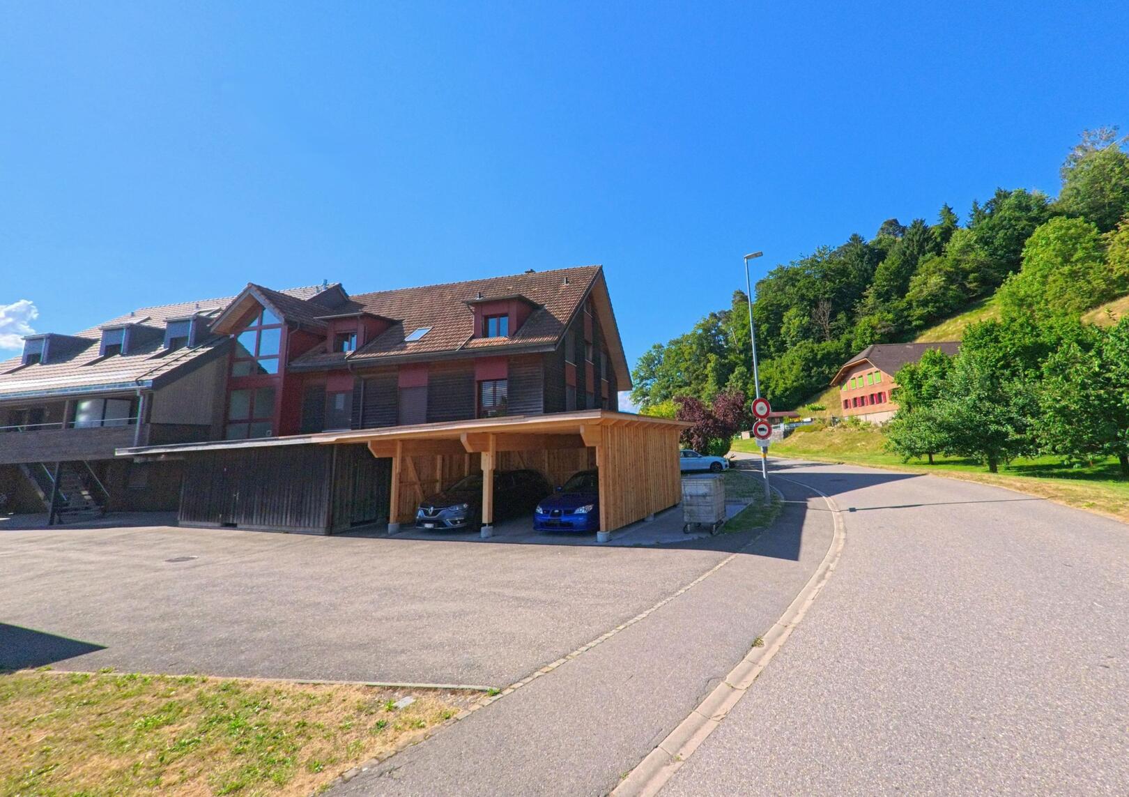 Am Rand von Oppligen (3629, Kanton Bern) verläuft eine ruhige Strasse entlang moderner Holzhäuser mit Carport, unterhalb eines bewaldeten Hangs mit einem roten Gebäude im Hintergrund.