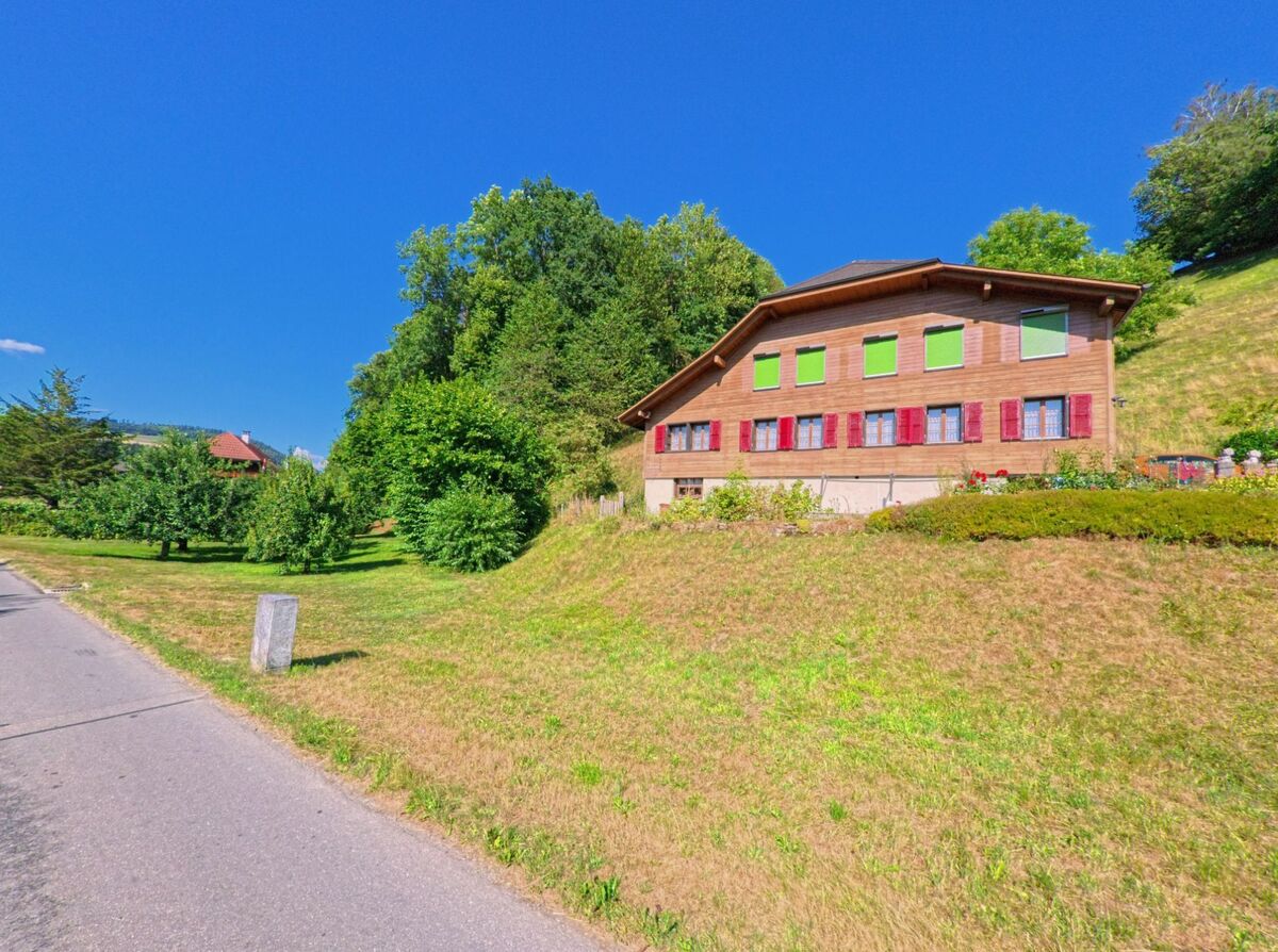 In Oppligen (3629, Kanton Bern) steht ein Holzhaus mit roten und grünen Fensterläden am Hang oberhalb einer gepflegten Obstwiese und wird von Bäumen und Wiesen umgeben.