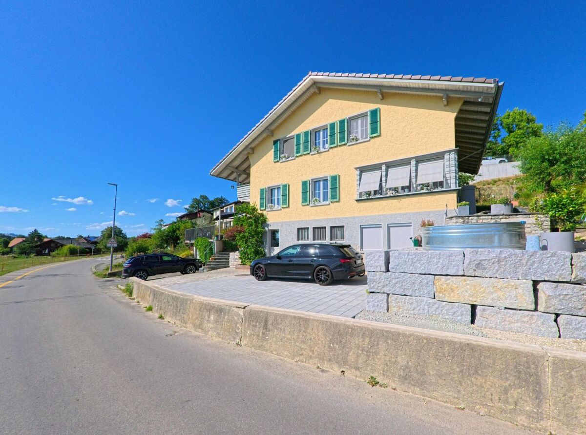 In Oppligen (3629, Kanton Bern) steht ein gelbes Wohnhaus mit grünen Fensterläden an einer leicht geschwungenen Strasse, flankiert von Gartenmauern und geparkten Autos.