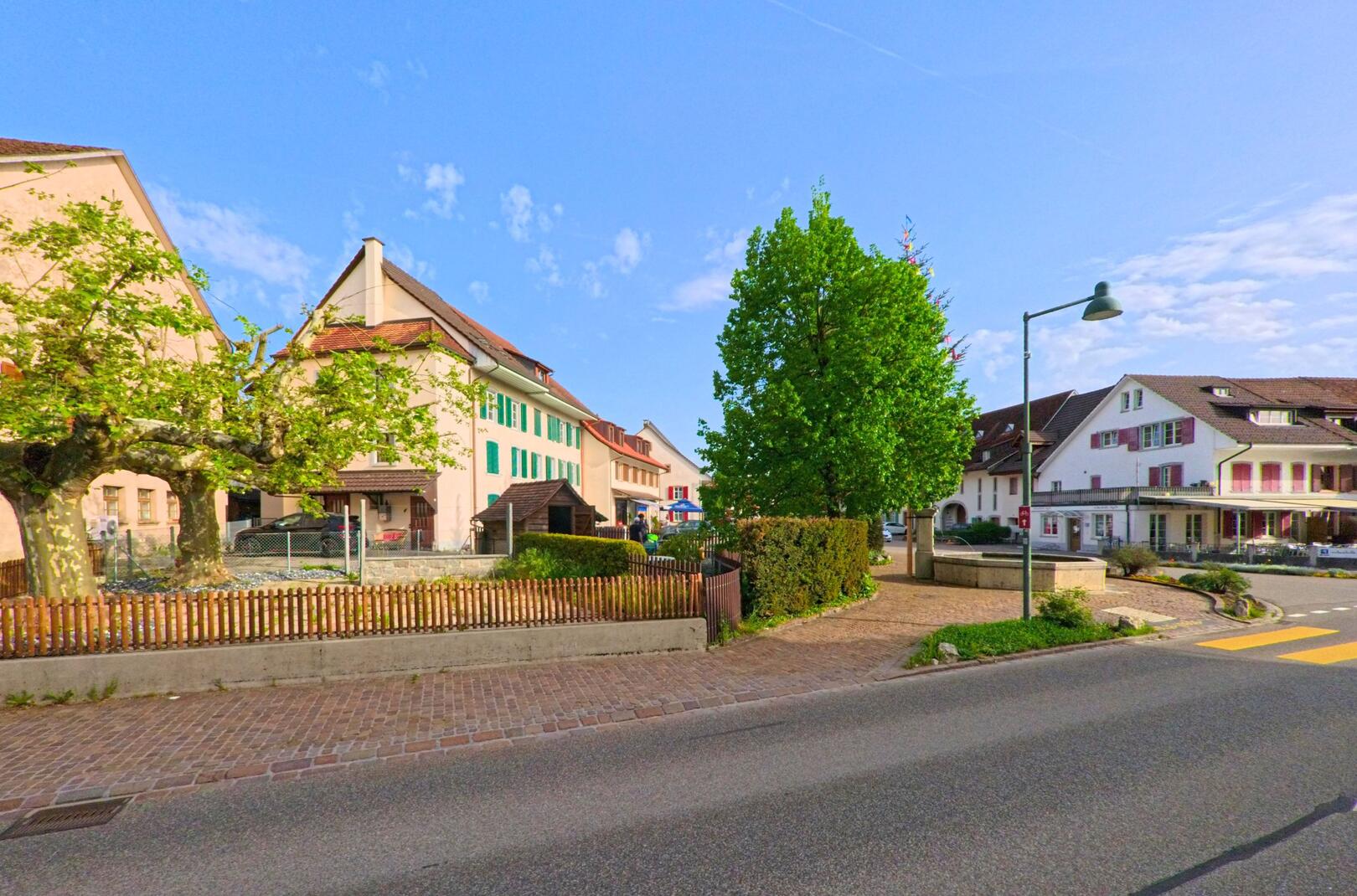 In Ormalingen (PLZ 4466, Kanton Baselland) säumen traditionelle Wohnhäuser mit Fensterläden und ein Baumplatz mit Brunnen eine verkehrsberuhigte Strassenecke im Dorfzentrum.
