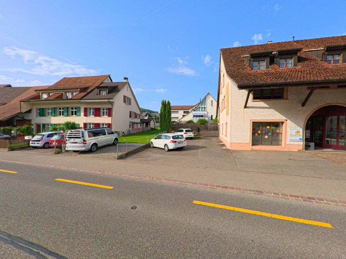 In Ormalingen (PLZ 4466, Kanton Baselland) liegen ein Kindergarten mit bemalten Fenstern und mehrere Wohnhäuser mit Parkplätzen entlang einer Hauptstrasse.