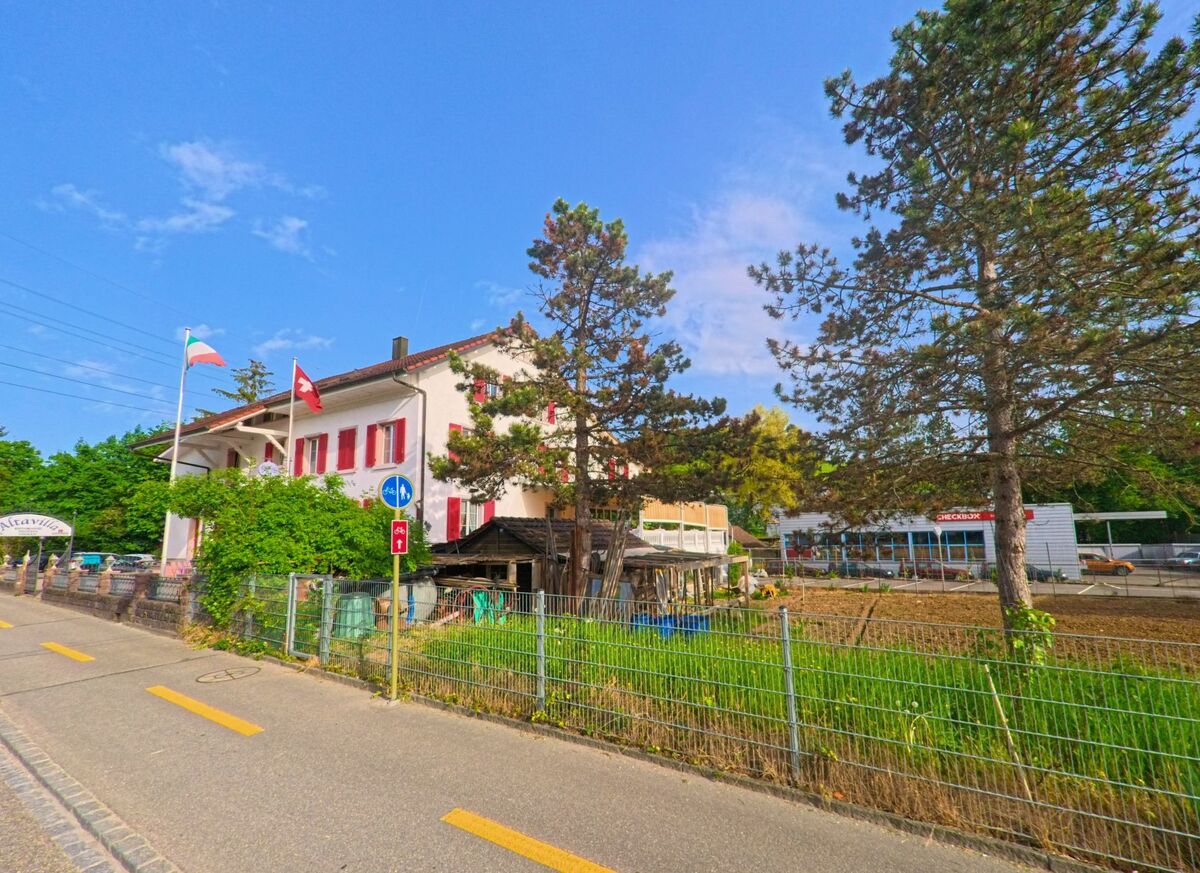In Ormalingen (PLZ 4466, Kanton Baselland) steht ein Haus mit roten Fensterläden und Schweizer Flagge direkt an einem Radweg neben einem eingezäunten Garten und Gewerbebauten.