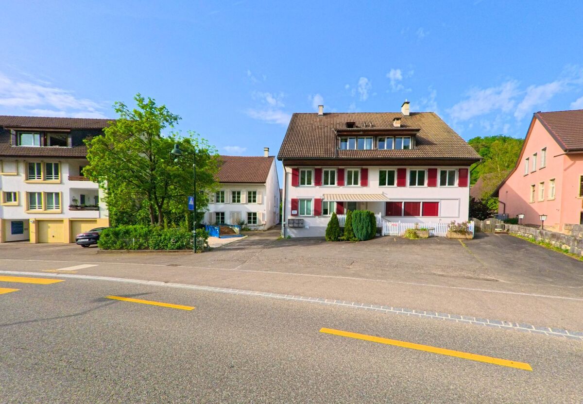 In Ormalingen (PLZ 4466, Kanton Baselland) stehen Wohnhäuser mit roten Fensterläden und Vorgärten entlang einer breiten Strasse mit Fussgängerübergang.