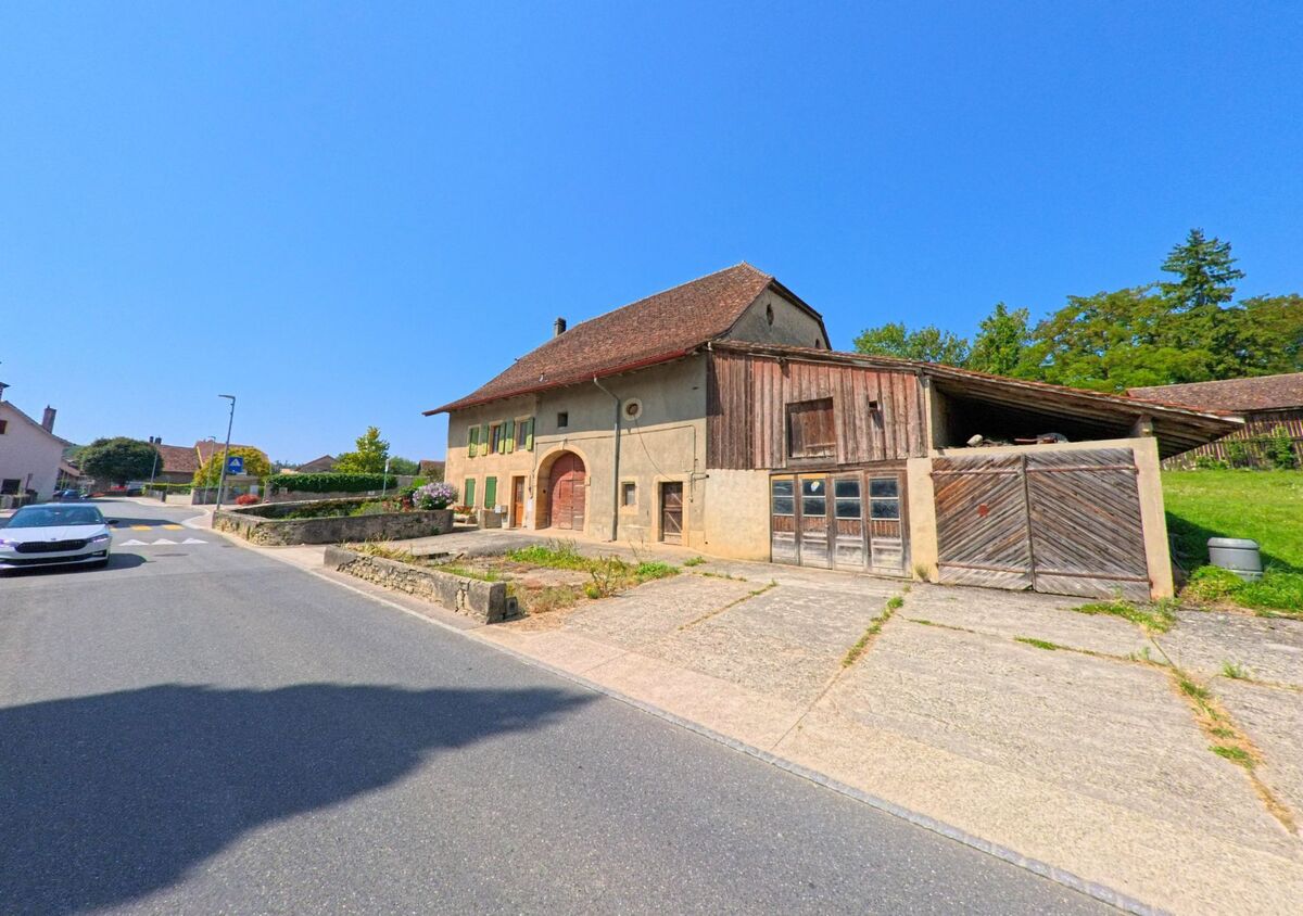 Ein traditionelles Bauernhaus mit Holzscheune und gruener Umgebung steht an einer Strasse in Orny (1317, Kanton Waadt).