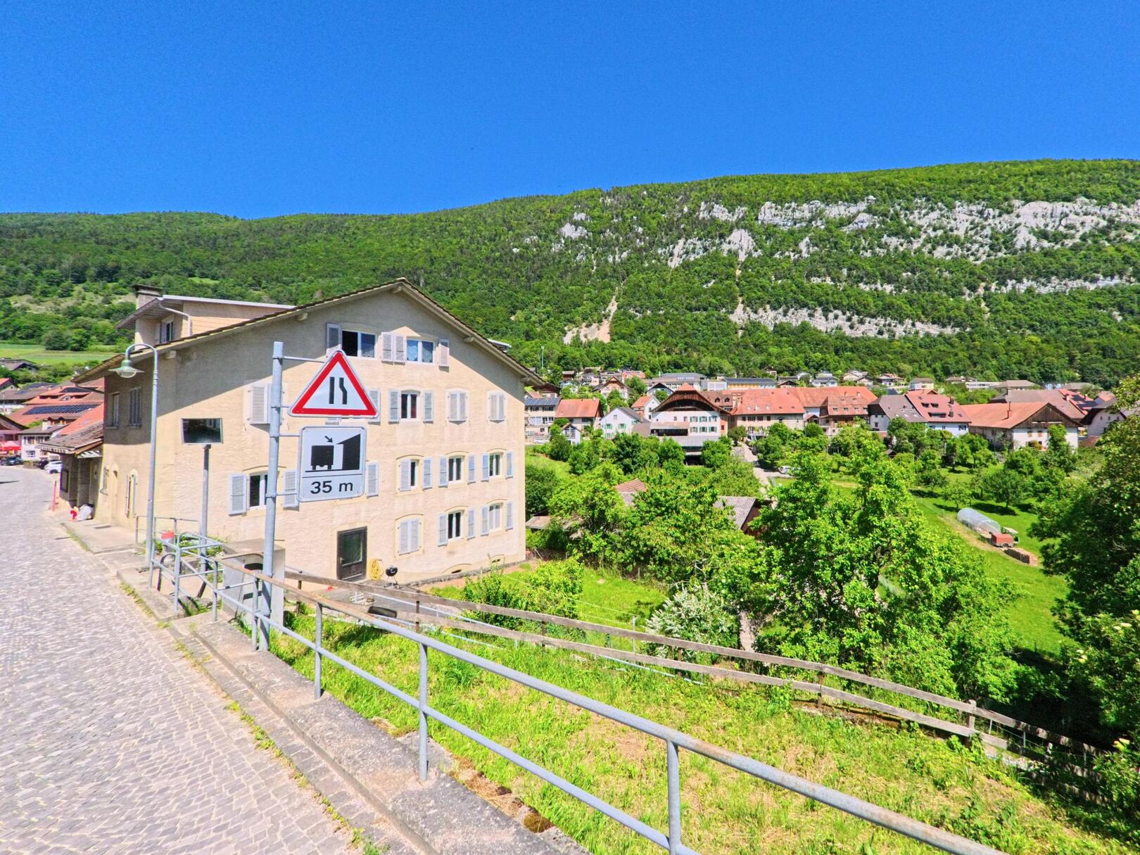 In Orvin (2534, Kanton Bern) verläuft eine gepflasterte Strasse an einem mehrstöckigen Gebäude vorbei, mit Blick auf das Dorf und die bewaldeten Jurahänge.