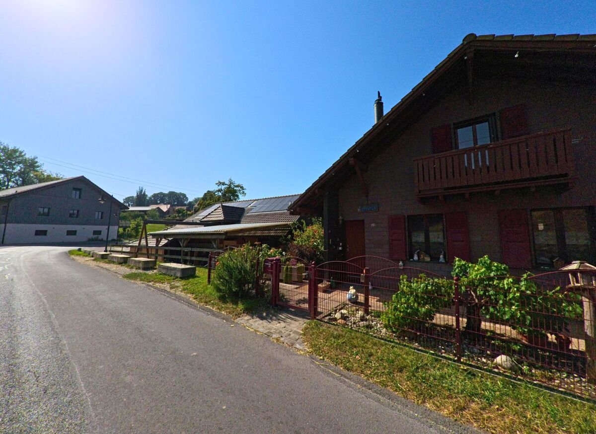 Ein Chalet mit roten Fensterlaeden und Balkon steht in Orzens (1413, Kanton Waadt) an einer ruhigen Strasse, nebenan befindet sich ein Nebengebaeude mit Solarpanelen.
