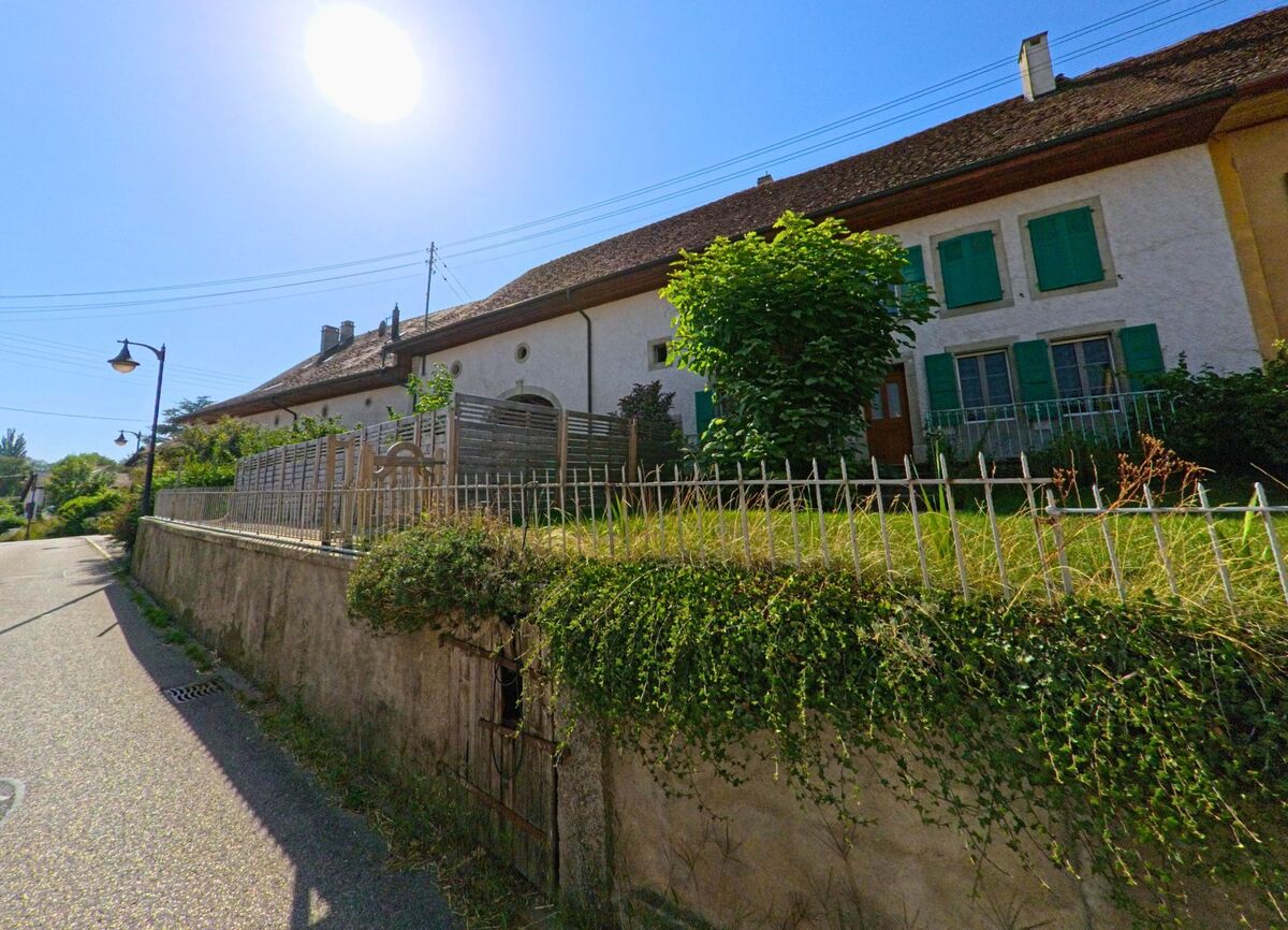 Ein langgestrecktes Wohnhaus mit gruenen Fensterlaeden in Orzens (1413, Kanton Waadt) steht oberhalb einer Mauer mit Bewuchs und einem eisernen Gittertor.