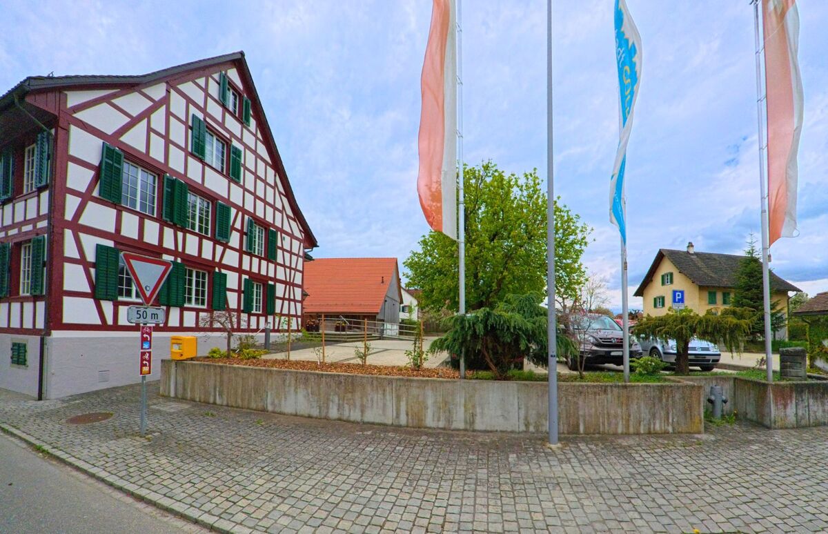 In Ossingen, PLZ 8475 im Kanton Zürich, steht ein auffällig gestaltetes Fachwerkhaus mit grünen Fensterläden direkt an einer gepflasterten Strasse mit Fahnenmasten. Bildtitel: ossingen8475_fachwerkhaus_fahnenplatz