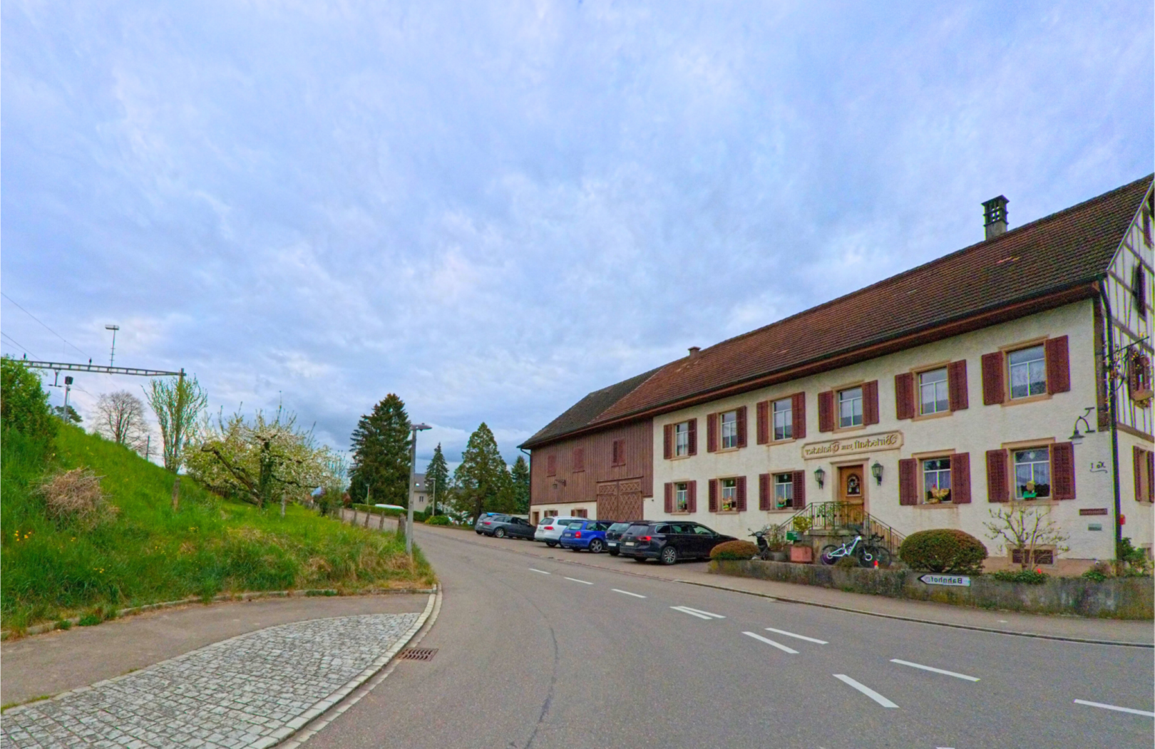 In Ossingen, PLZ 8475 im Kanton Zürich, steht ein traditionelles Gasthaus mit Fensterläden entlang einer leicht geschwungenen Dorfstrasse gegenüber einer begrünten Böschung.