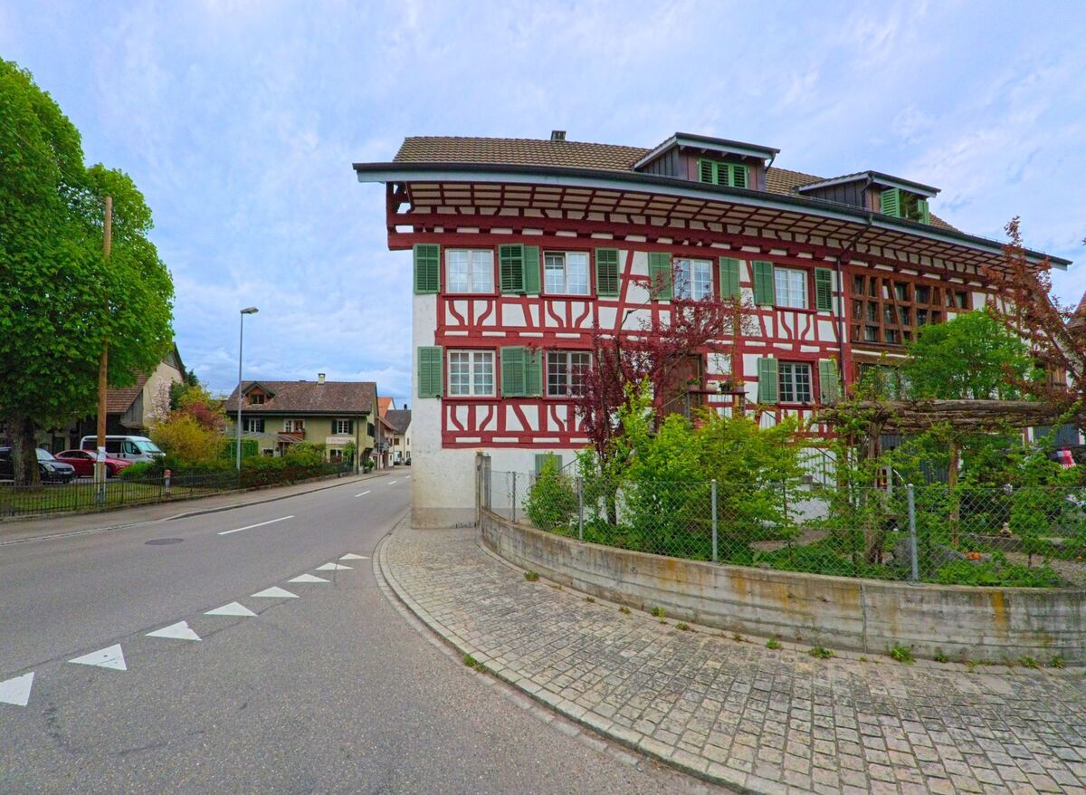 In Ossingen, PLZ 8475 im Kanton Zürich, verläuft eine Hauptstrasse an einem imposanten Fachwerkhaus mit gruenen Fensterläden und Vorgarten entlang.