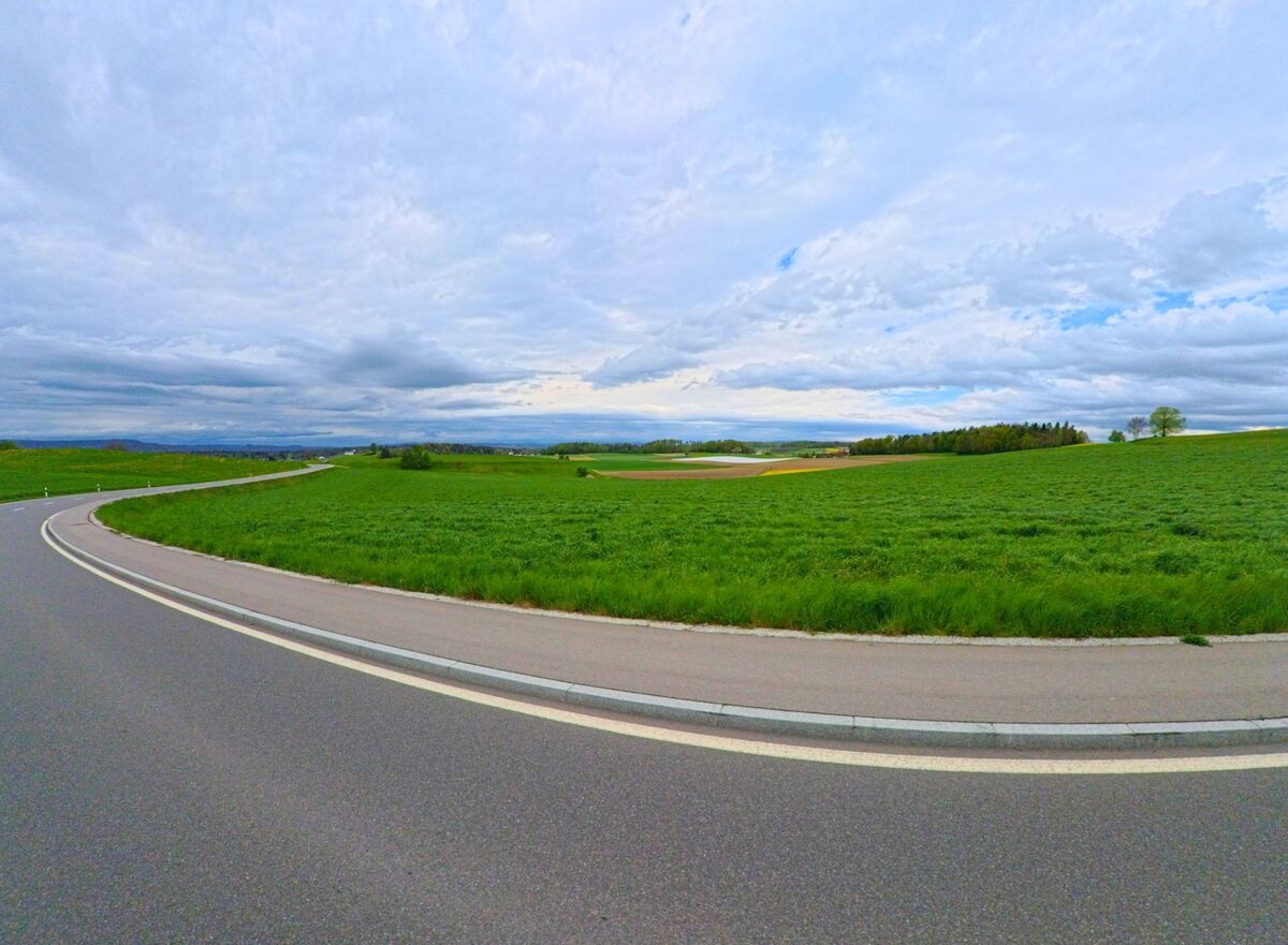 In Ossingen, PLZ 8475 im Kanton Zürich, schlängelt sich eine Landstrasse durch weitläufige Felder mit Blick auf bewaldete Hügel in der Ferne.