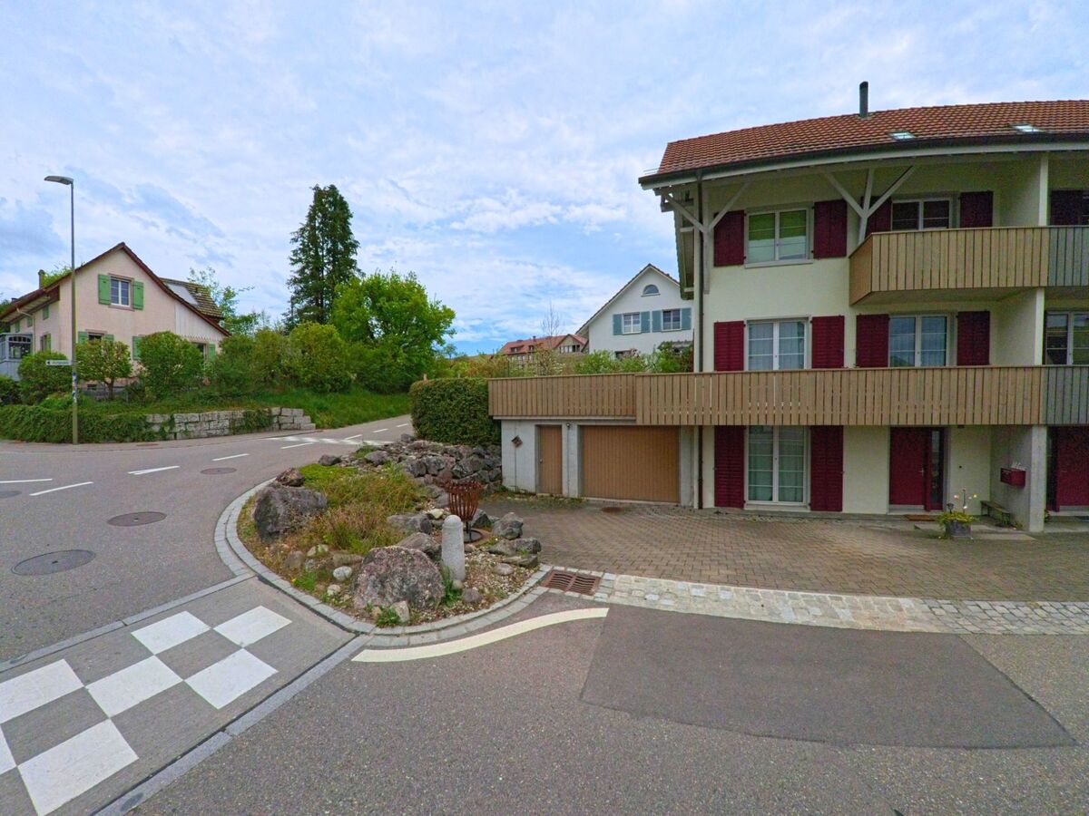 In Ossingen, PLZ 8475 im Kanton Zürich, treffen moderne Wohnhäuser mit Balkonen auf eine verkehrsberuhigte Strassenecke mit begrüntem Randbereich.