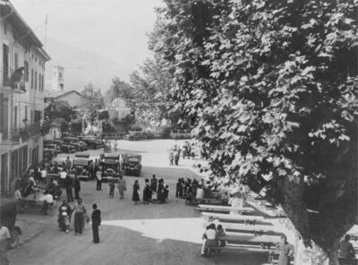 Eine historische Aufnahme aus Caslano (6987, Kanton Tessin) zeigt Menschen und Autos auf einem Platz, umgeben von grossen Bäumen und Gebäuden.