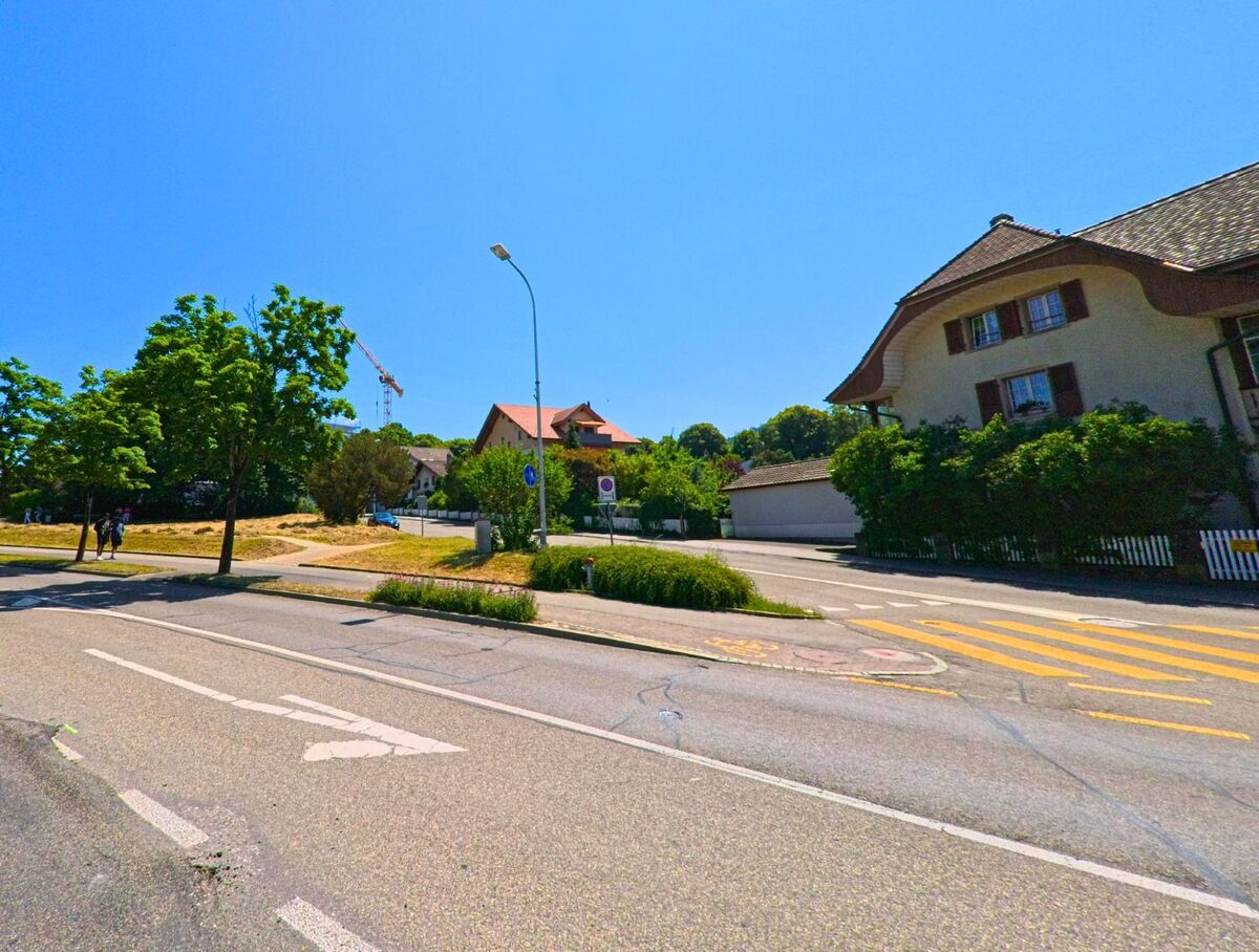 In Ostermundigen (3072, Kanton Bern) führt eine Straße an einer Baumreihe entlang, während im Hintergrund ein Gebäude und ein Baukran sichtbar sind.