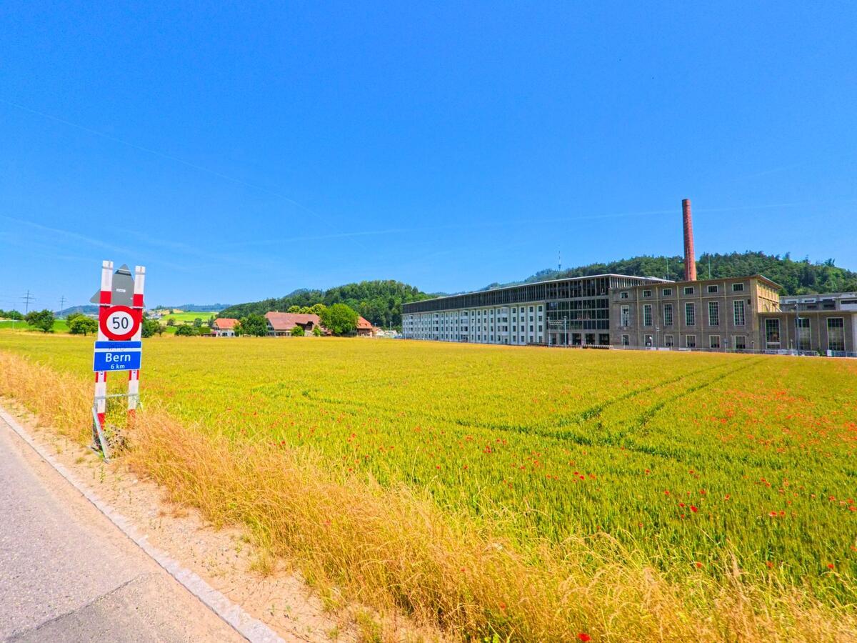 In Ostermundigen (3072, Kanton Bern) führt eine Straße vorbei an einem weitläufigen Feld und einem ehemaligen Industriebau mit einem hohen Schornstein.