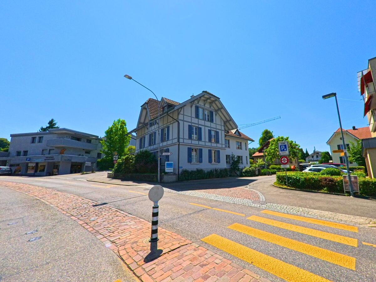 In Ostermundigen (3072, Kanton Bern) führt eine Straße an einem historischen Gebäude und modernen Wohnhäusern entlang, während ein Zebrastreifen und Verkehrsschilder zu sehen sind.