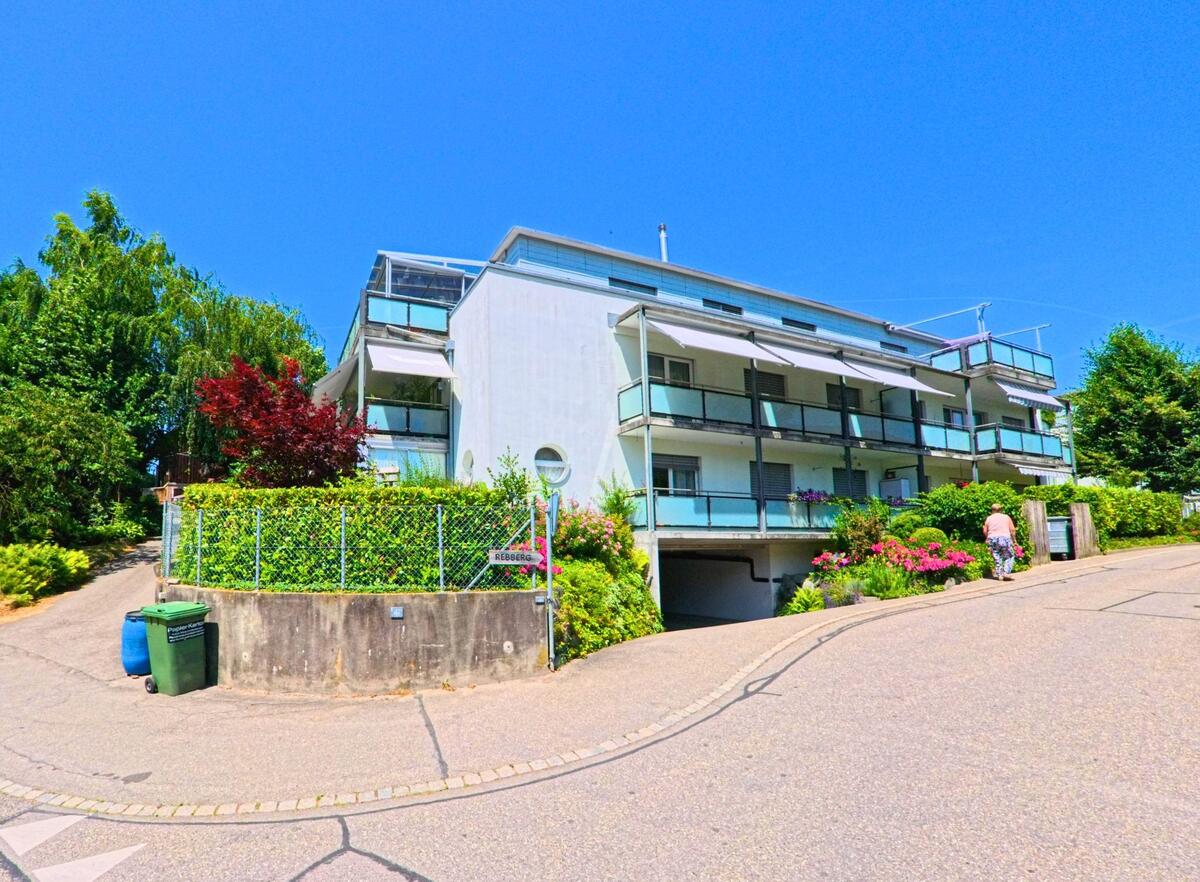 In Ostermundigen (3072, Kanton Bern) führt eine leicht ansteigende Straße an einem modernen Gebäude mit Balkonen und einem gepflegten Garten entlang.