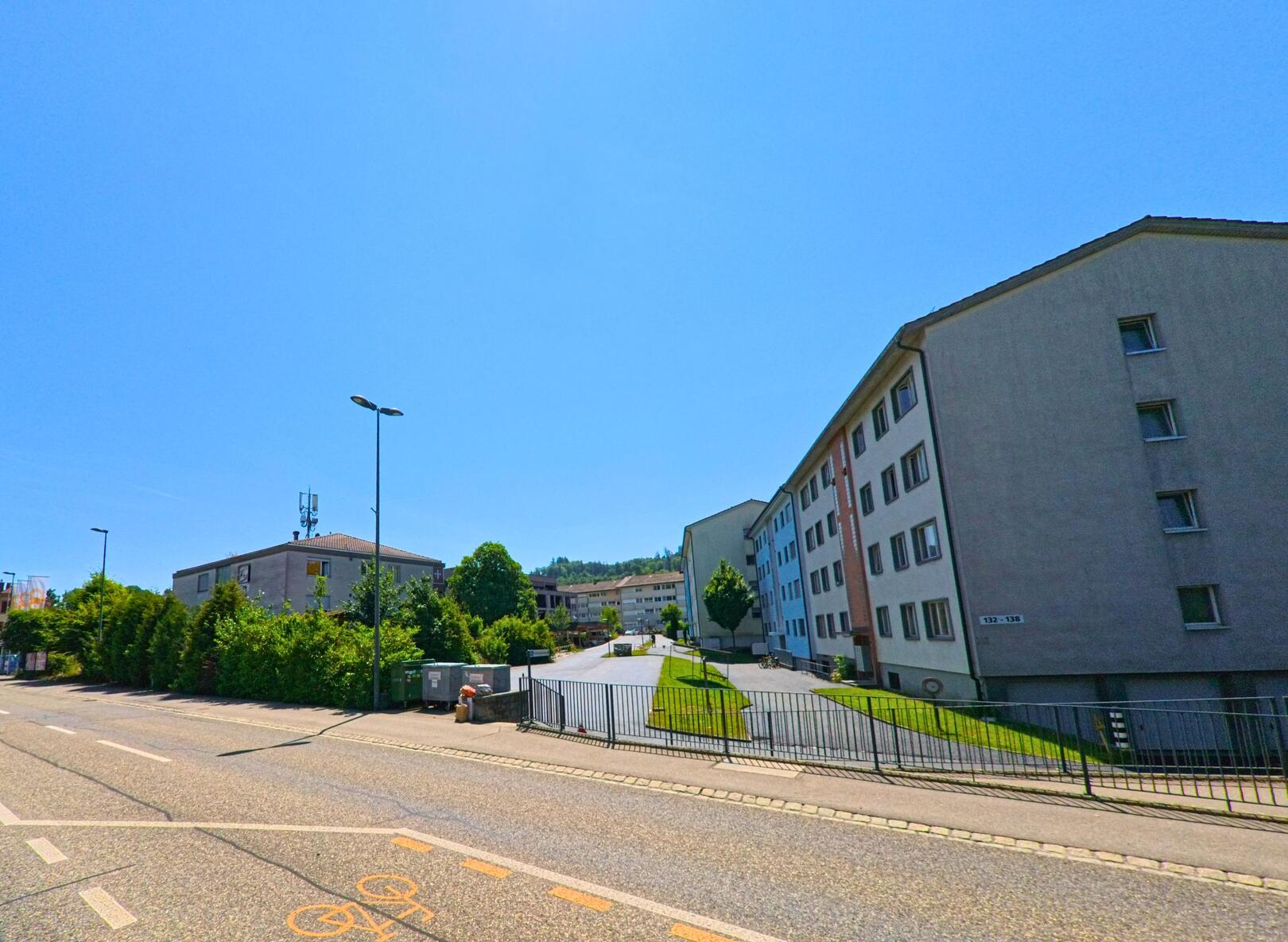 In Ostermundigen (3072, Kanton Bern) erstreckt sich eine Strasse entlang von Wohngebäuden, während ein Motorradfahrer die Strecke bei sonnigem Wetter fährt.