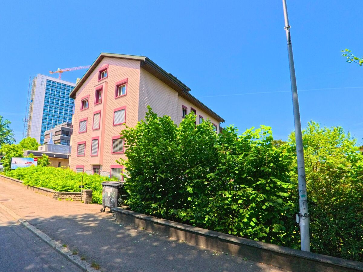 In Ostermundigen (3072, Kanton Bern) ist eine moderne Bürogebäudekonstruktion im Hintergrund sichtbar, während ein Wohngebäude und viel Grün entlang der Straße im Vordergrund stehen.