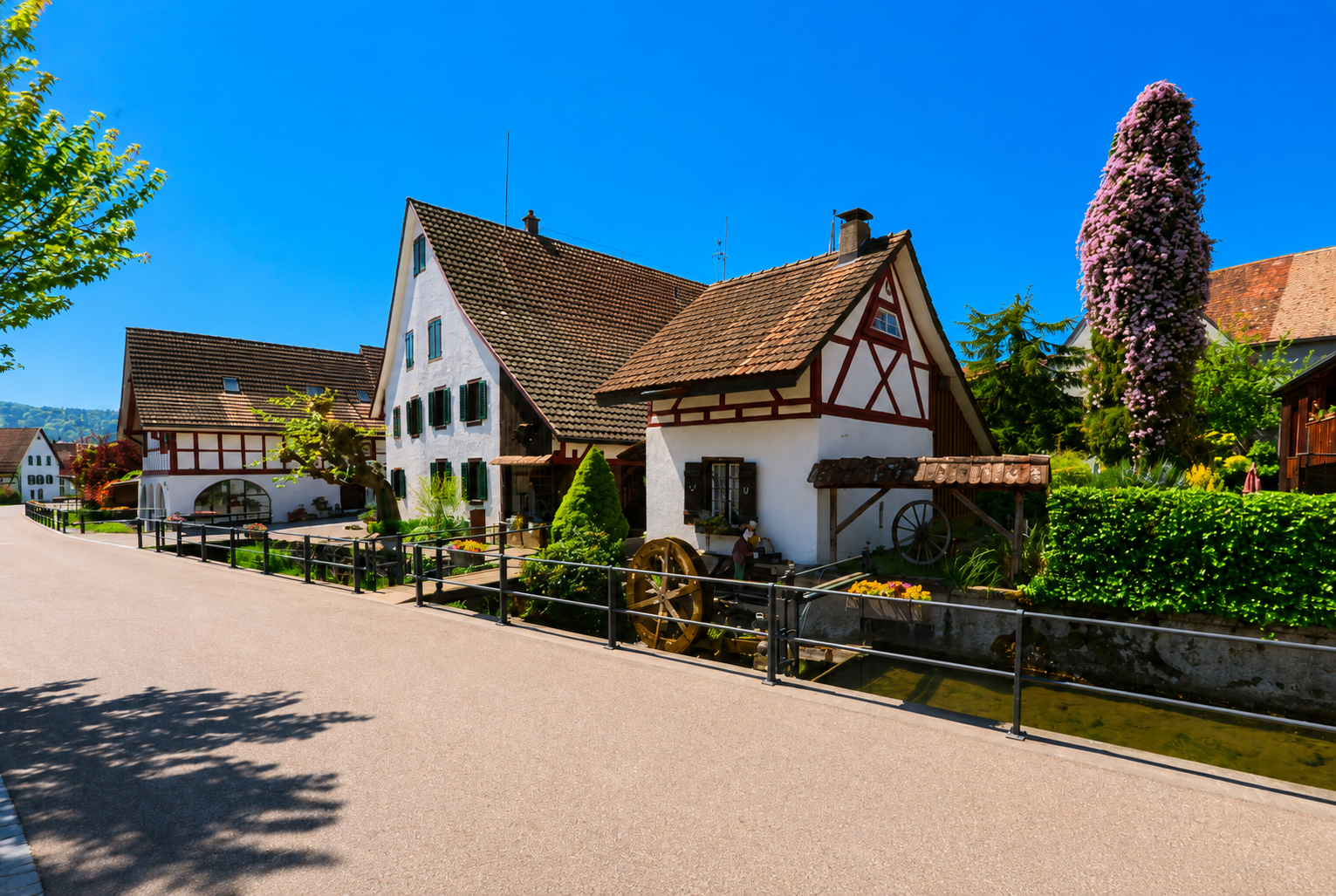 Entlang einer Strasse in Otelfingen (8112, Kanton Zürich) stehen traditionelle Fachwerkhäuser neben einem schmalen Wasserlauf unter blauem Himmel.