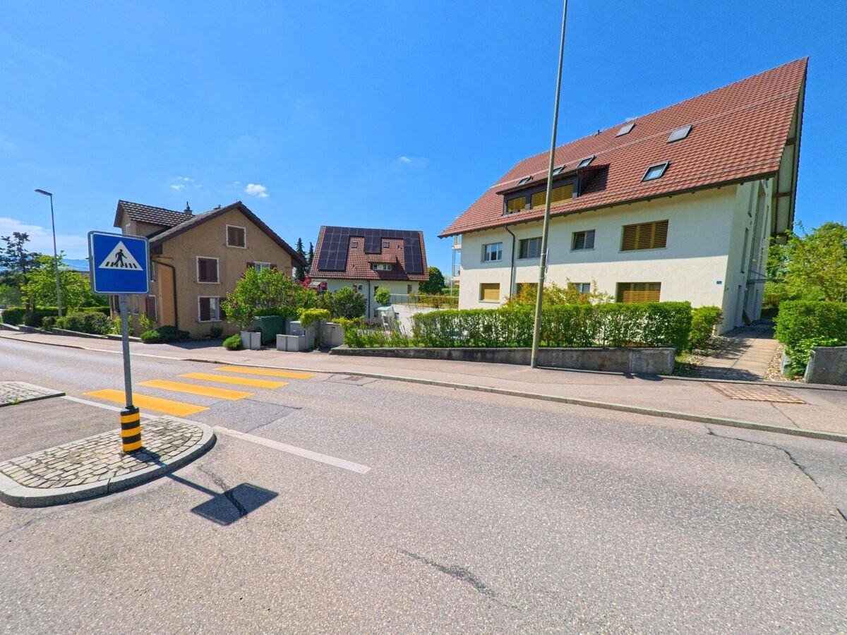 Ein Fussgängerstreifen in Ottenbach im Kanton Zürich mit der PLZ 8913 führt über eine Quartierstrasse, flankiert von Wohnhäusern mit gepflegten Vorgärten und Solarpanels.