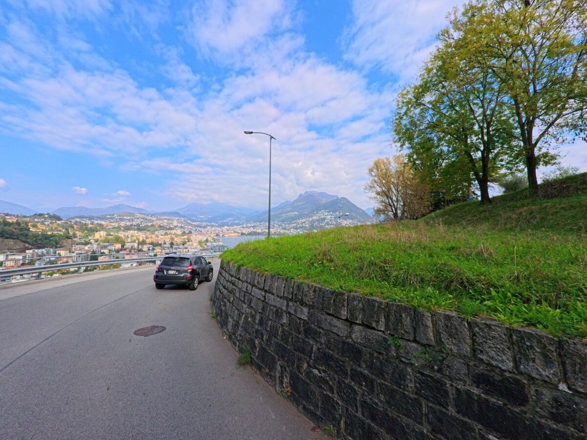 Entlang einer Hangstrasse mit Steinmauer eröffnet sich in Paradiso (6900, Kanton Tessin) der Blick auf die Stadt, den See und die umliegenden Berge unter wechselnd bewölktem Himmel.
