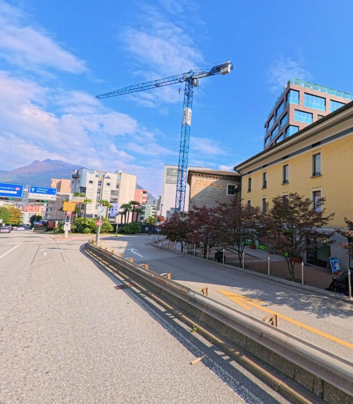 An einer Hauptstrasse mit Leitplanken zeigt sich in Paradiso (6900, Kanton Tessin) ein städtischer Bereich mit Baukran, modernen Gebäuden und Blick auf die umliegenden Berge.