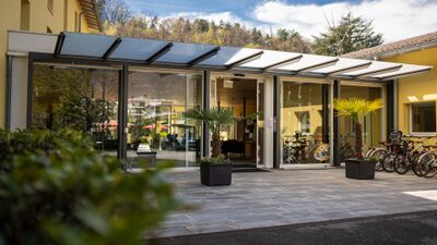 Der Eingang des Parkhotel Emmaus in Losone (6616, Kanton Tessin) ist von Glasfronten, Palmen und Fahrrädern gesäumt und führt in eine helle Empfangshalle.