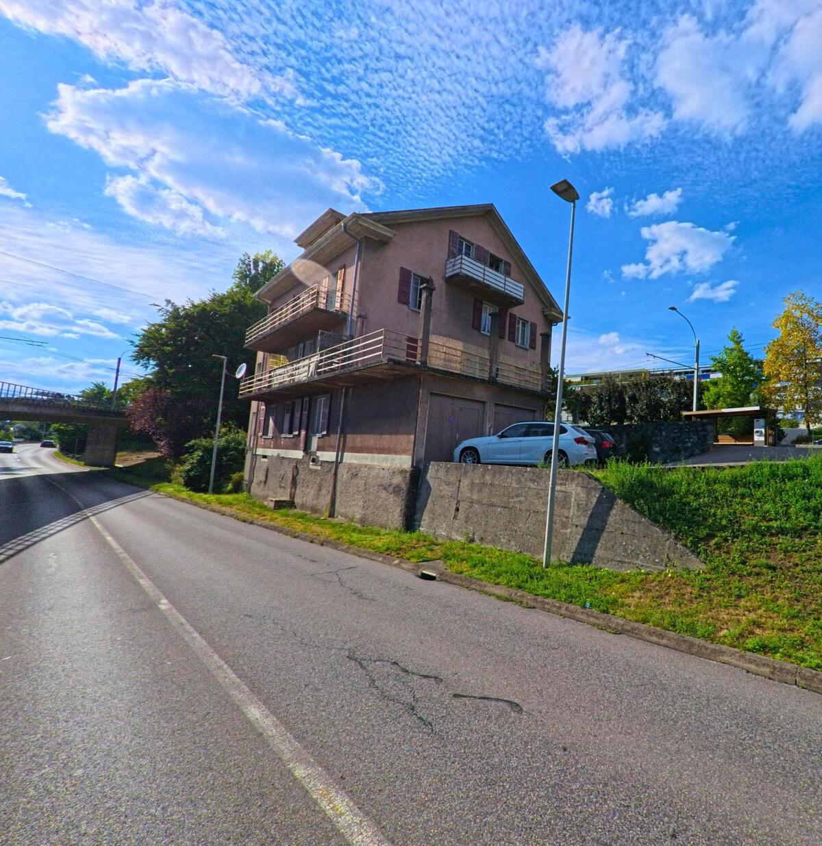 Ein aelteres Wohnhaus mit Balkonen steht an einer Strasse in Paudex (1094, Kanton Waadt), im Hintergrund ist eine Bruecke ueber die Fahrbahn zu sehen.