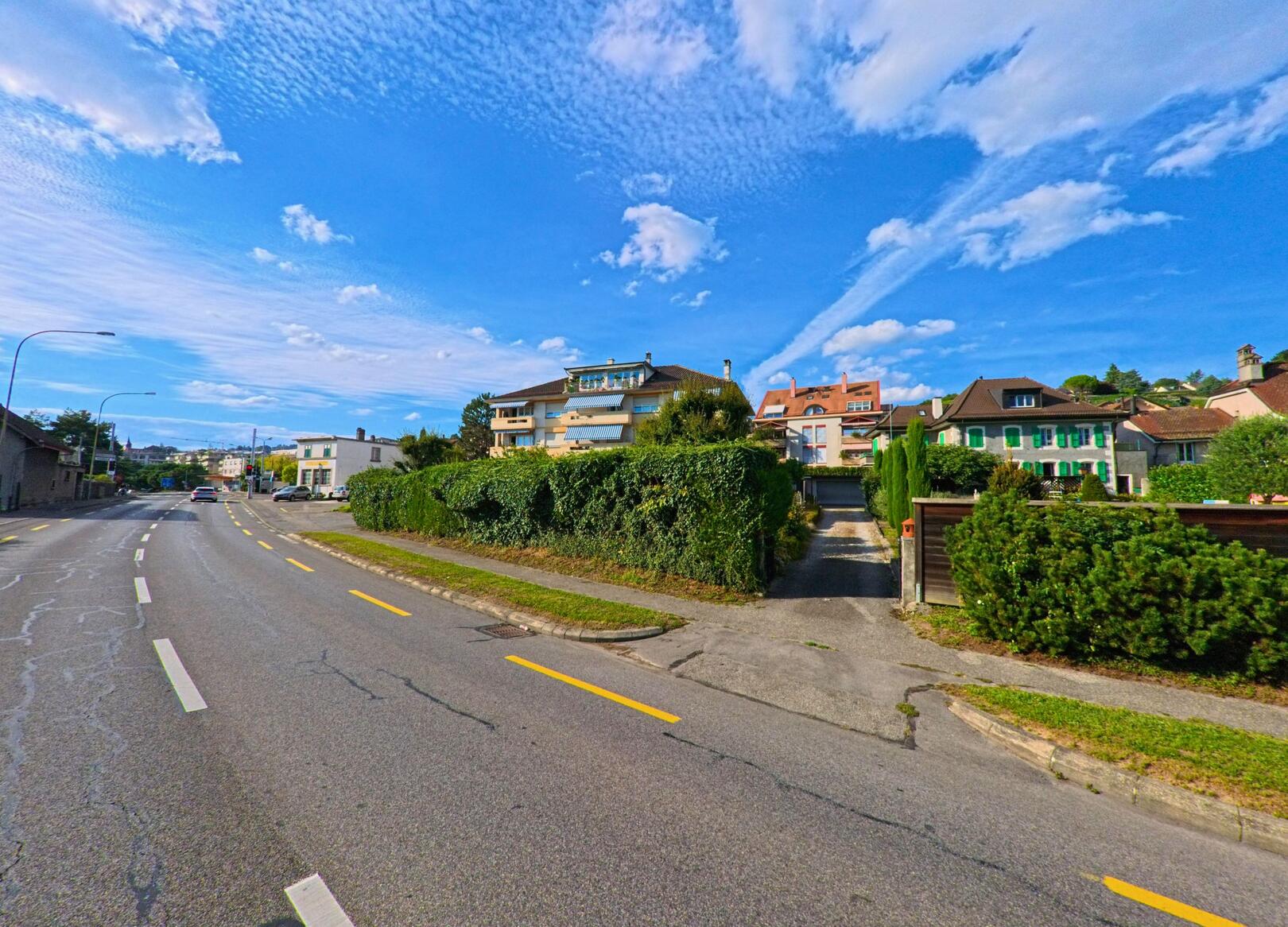 Entlang der Hauptstrasse in Paudex (1094, Kanton Waadt) stehen Wohnhaeuser mit gepflegten Gaerten, waehrend der Himmel von lockeren Wolken bedeckt ist.