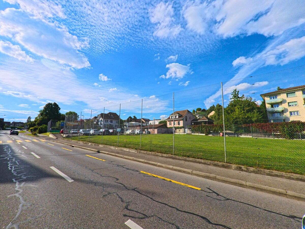 Ein umzäunter Rasenplatz liegt direkt an einer Strasse in Paudex (1094, Kanton Waadt), im Hintergrund sind Wohnhaeuser und parkende Autos zu sehen.