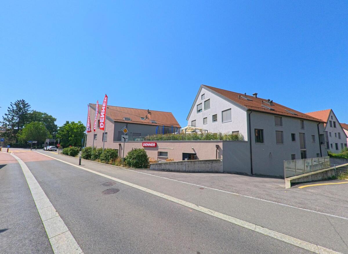 Ein modernes Wohnhaus mit rotem Dach in Penthaz (1303, Kanton Waadt) beherbergt im Erdgeschoss eine Denner-Filiale mit auffaelligen Fahnen.