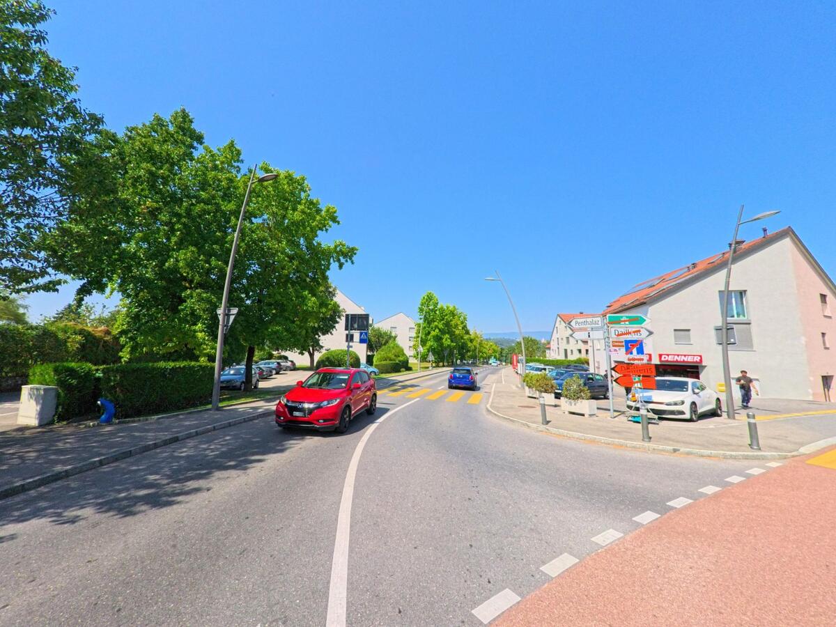 Eine Strasse mit Fussgaengerstreifen fuehrt in Penthaz (1303, Kanton Waadt) an Wohnhaeusern und einem Denner-Laden vorbei.