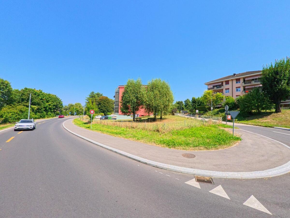 Eine geschwungene Strasse mit angrenzenden Wohnblocks und Gruenflaechen zeigt eine typische Szene in Penthaz (1303, Kanton Waadt).