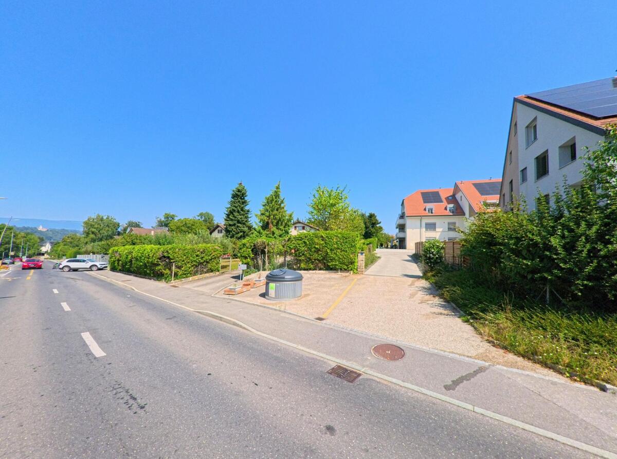 Am Strassenrand in Penthaz (1303, Kanton Waadt) liegen moderne Wohnhaeuser mit roten Daechern, daneben ein kleiner Parkplatz mit Abfallstation.
