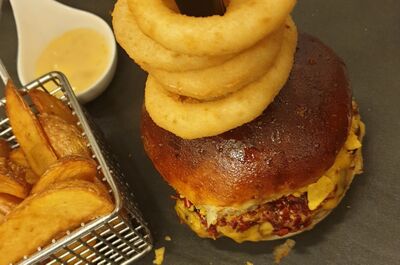 Im Pepper Grill in Melano (6818, Kanton Tessin) wird ein Burger mit knusprigen Onion Rings, Kartoffelwedges und Dip-Sauce auf einem modernen Serviertablett präsentiert.