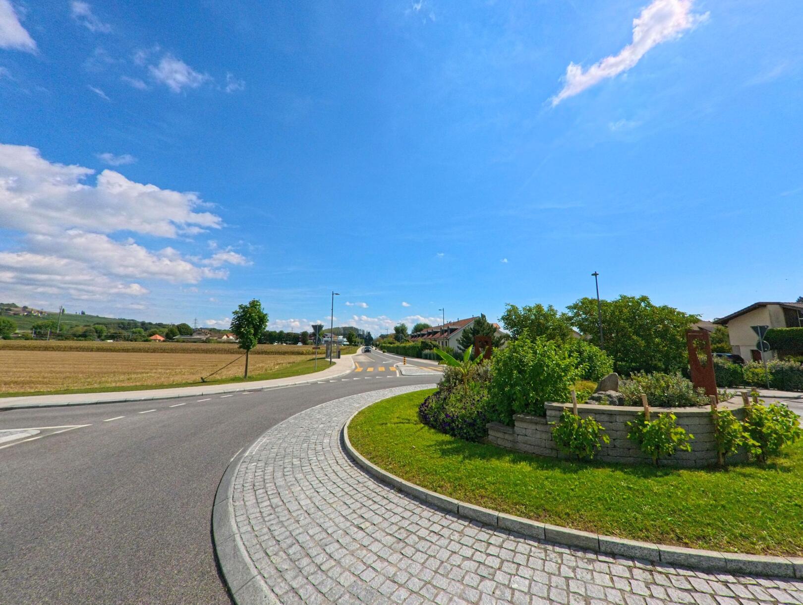 Ein begrünter Kreisverkehr in Perroy (1166, Kanton Waadt) verbindet die Strasse zwischen Feldern und Wohnhäusern bei klarem Himmel.