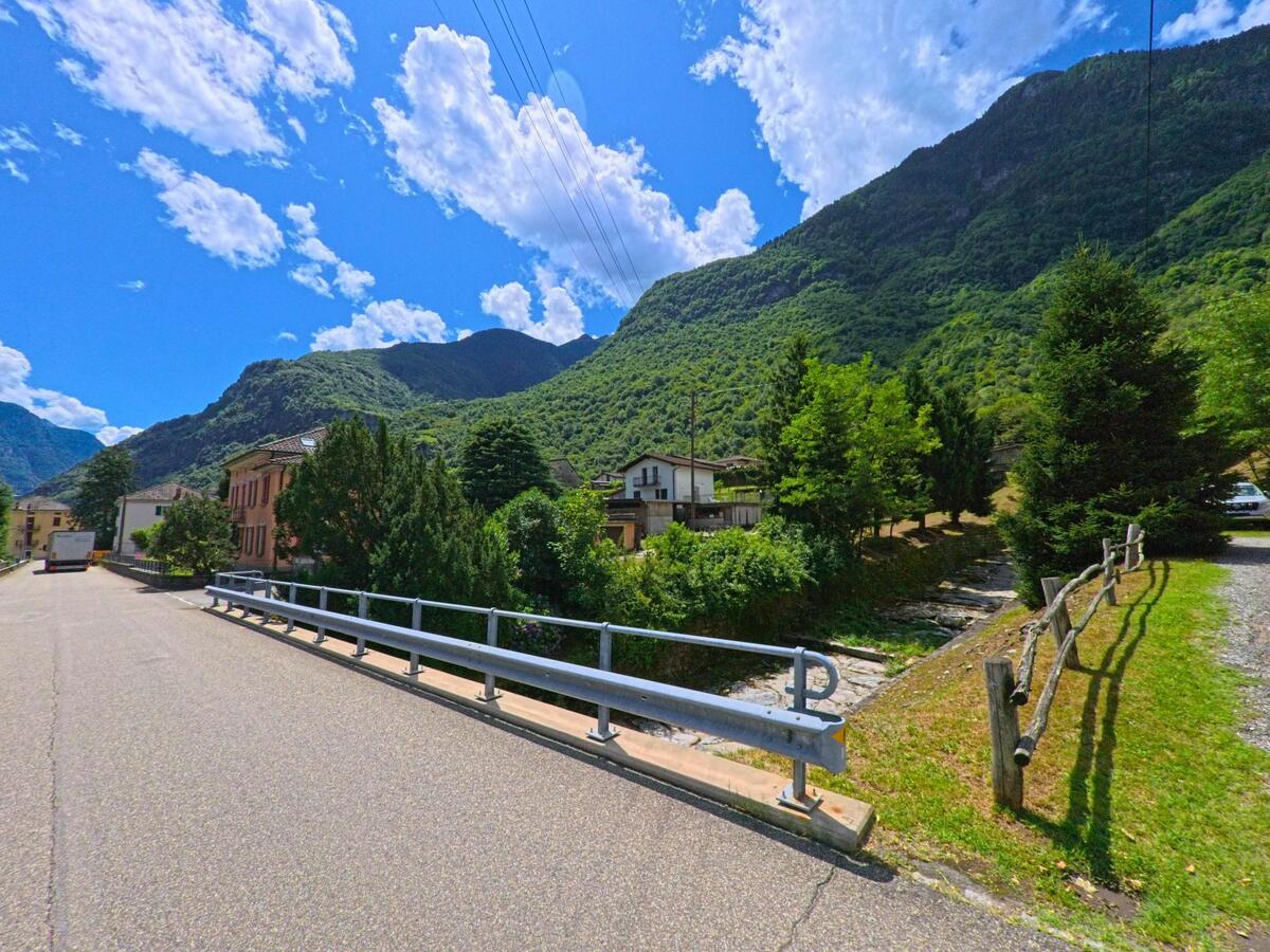 Eine Strasse in Personico (6744, Kanton Tessin) führt über eine Brücke mit Geländer entlang eines kleinen Baches, eingerahmt von bewaldeten Bergen.