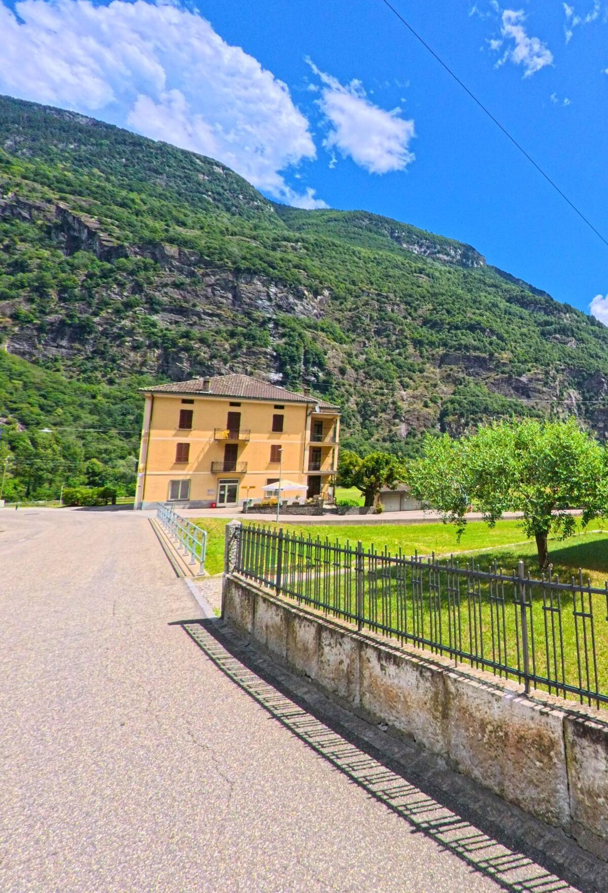 Ein gelbes Wohnhaus in Personico (6744, Kanton Tessin) steht am Rand einer ruhigen Strasse vor der steilen, grünen Bergflanke.