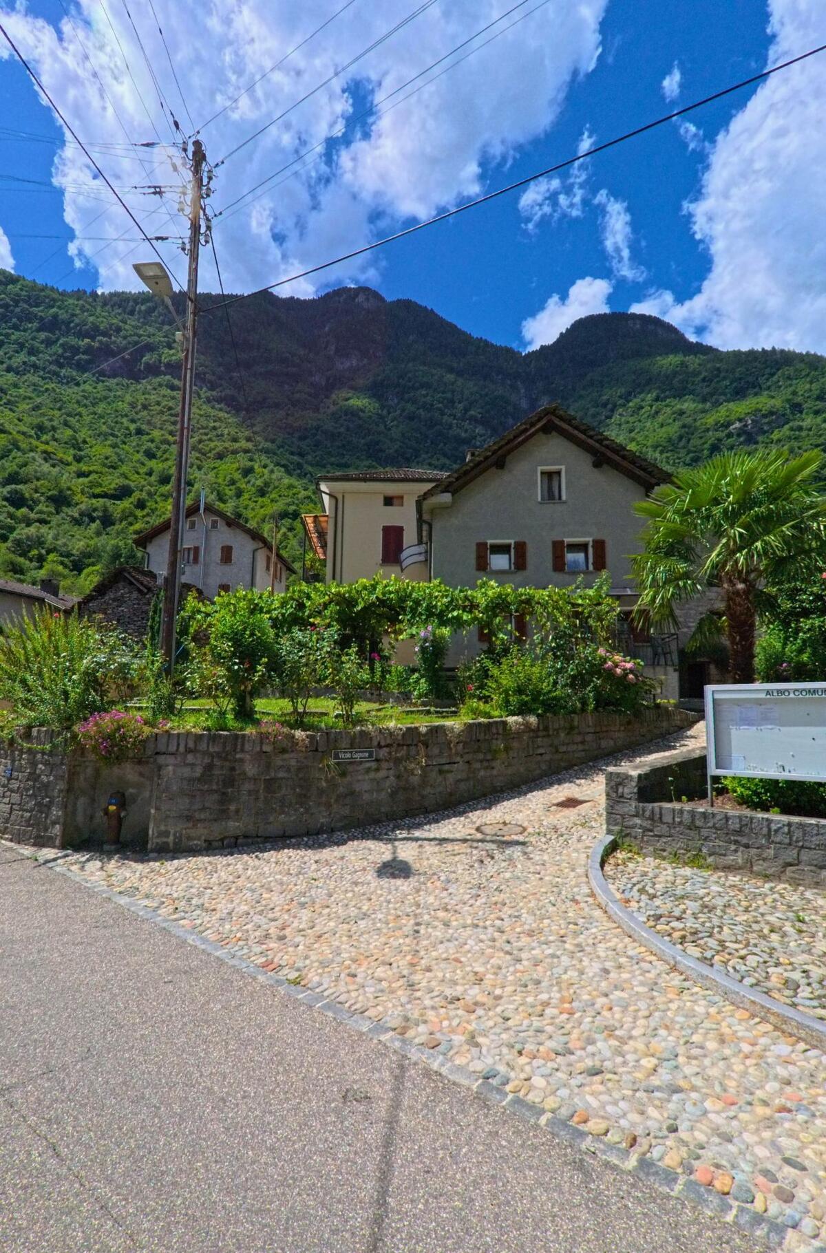 In Personico (6744, Kanton Tessin) führt ein gepflasterter Weg an gepflegten Gärten und traditionellen Häusern mit roten Fensterläden vorbei.