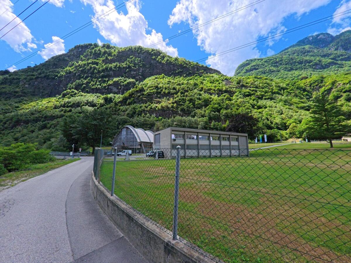 Am Ortsrand von Personico (6744, Kanton Tessin) steht eine moderne Halle mit umzäuntem Rasenplatz vor einer steilen, dicht bewaldeten Bergwand.