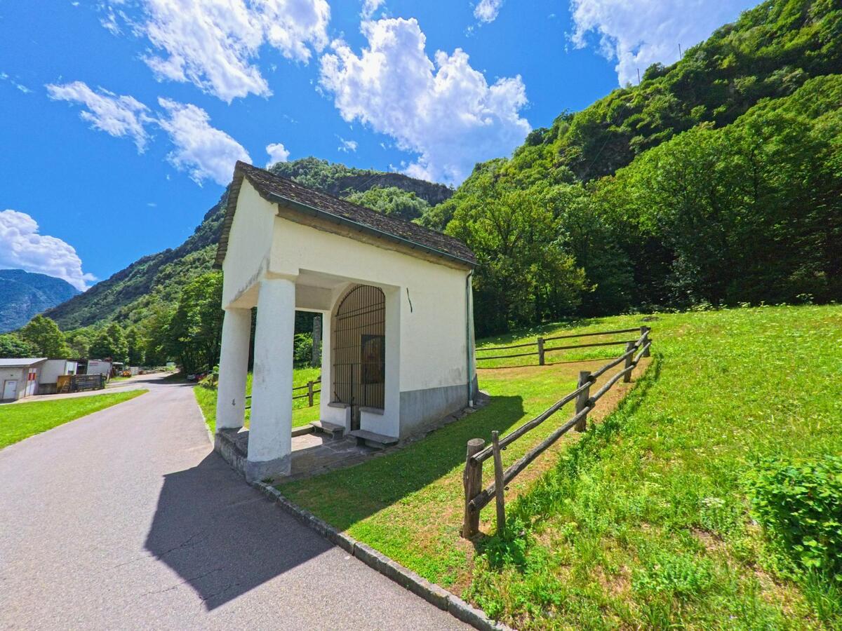 Am Strassenrand in Personico (6744, Kanton Tessin) steht eine kleine weisse Kapelle mit Gittertor vor einer grünen Wiese und bewaldeten Hügeln.