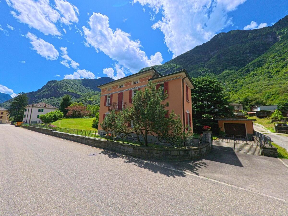 Ein rosafarbenes Haus mit roten Fensterläden steht in Personico (6744, Kanton Tessin) an einer ruhigen Strasse vor einer grünen Bergkulisse.