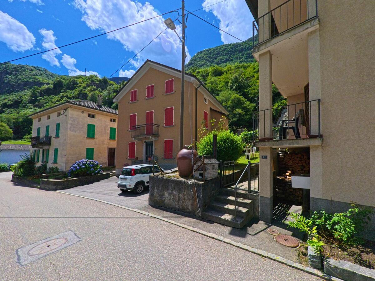 Mehrere Wohnhäuser mit bunten Fensterläden stehen in Personico (6744, Kanton Tessin) an einer engen Strasse am Fusse bewaldeter Hügel.