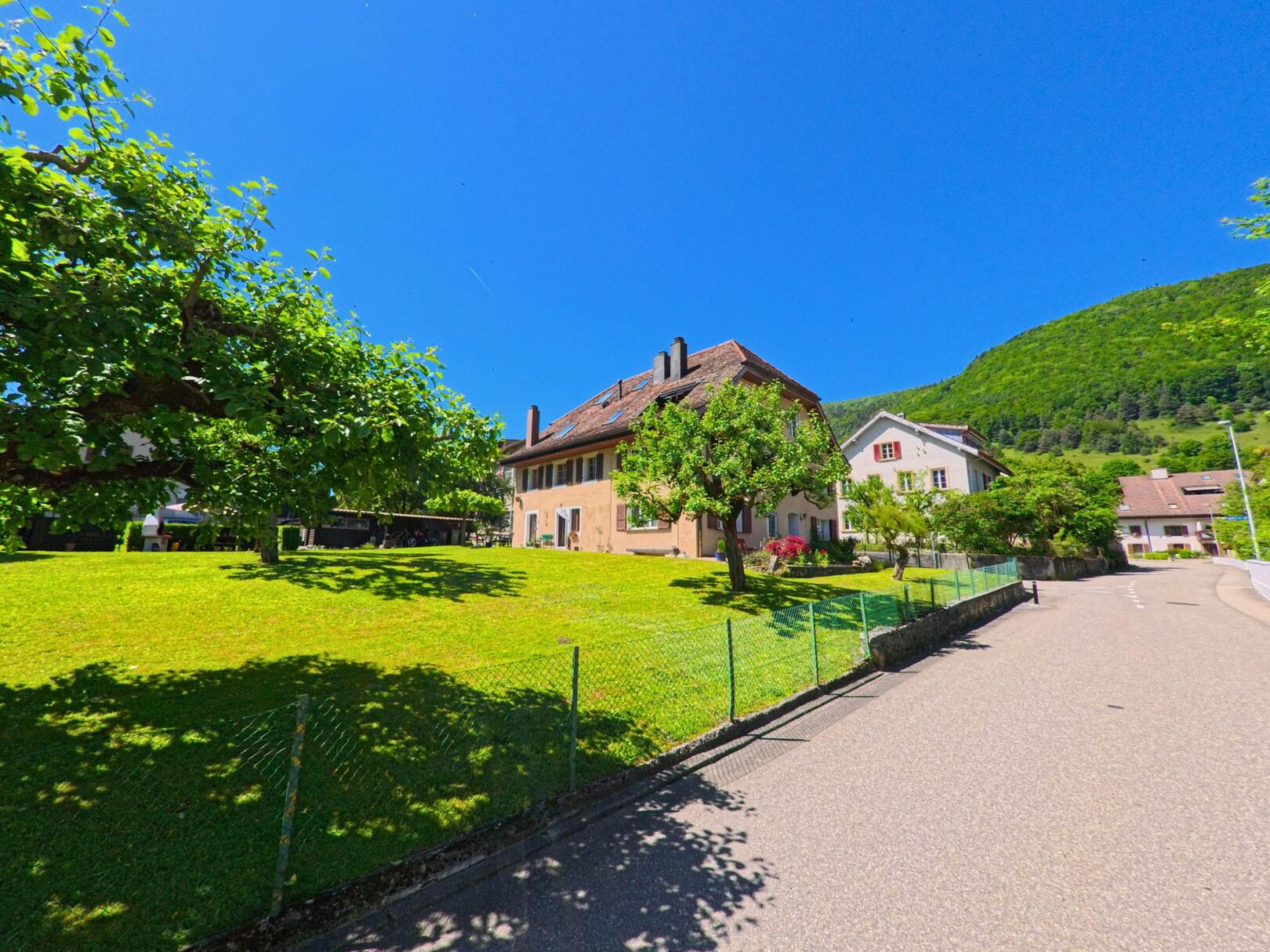 In Péry-La Heutte (2603, Kanton Bern) stehen mehrere Wohnhäuser mit grossen Gärten entlang einer ruhigen Strasse am grünen Hang.