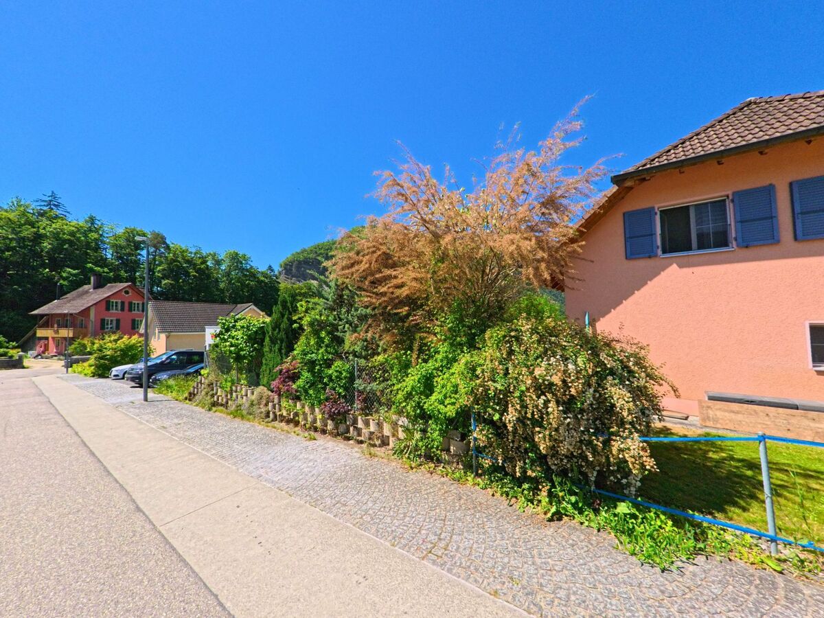 In Péry-La Heutte (2603, Kanton Bern) verläuft eine ruhige Straße mit Vorgärten und farbenfrohen Häusern entlang eines bewaldeten Hangs.